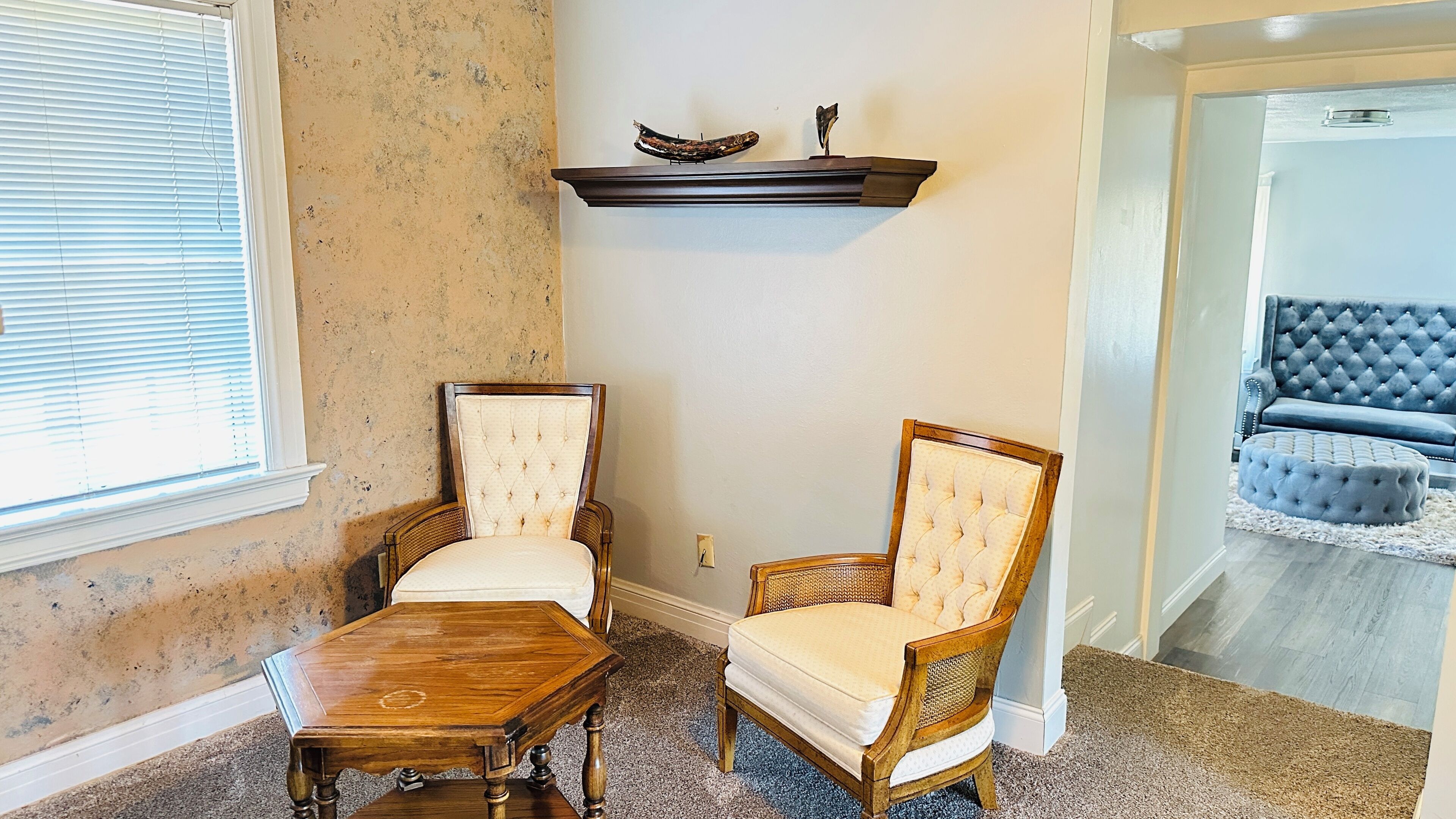 A corner room with two upholstered chairs, a wooden table, and a dark wooden shelf above.