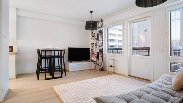 Un salon moderne comprenant un canapé, une table à manger avec des chaises, un téléviseur à écran plat et une grande fenêtre donnant sur des immeubles.