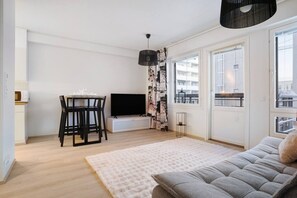 Een moderne woonkamer met een bank, een eettafel met stoelen, een flatscreen televisie en een groot raam met uitzicht op gebouwen.