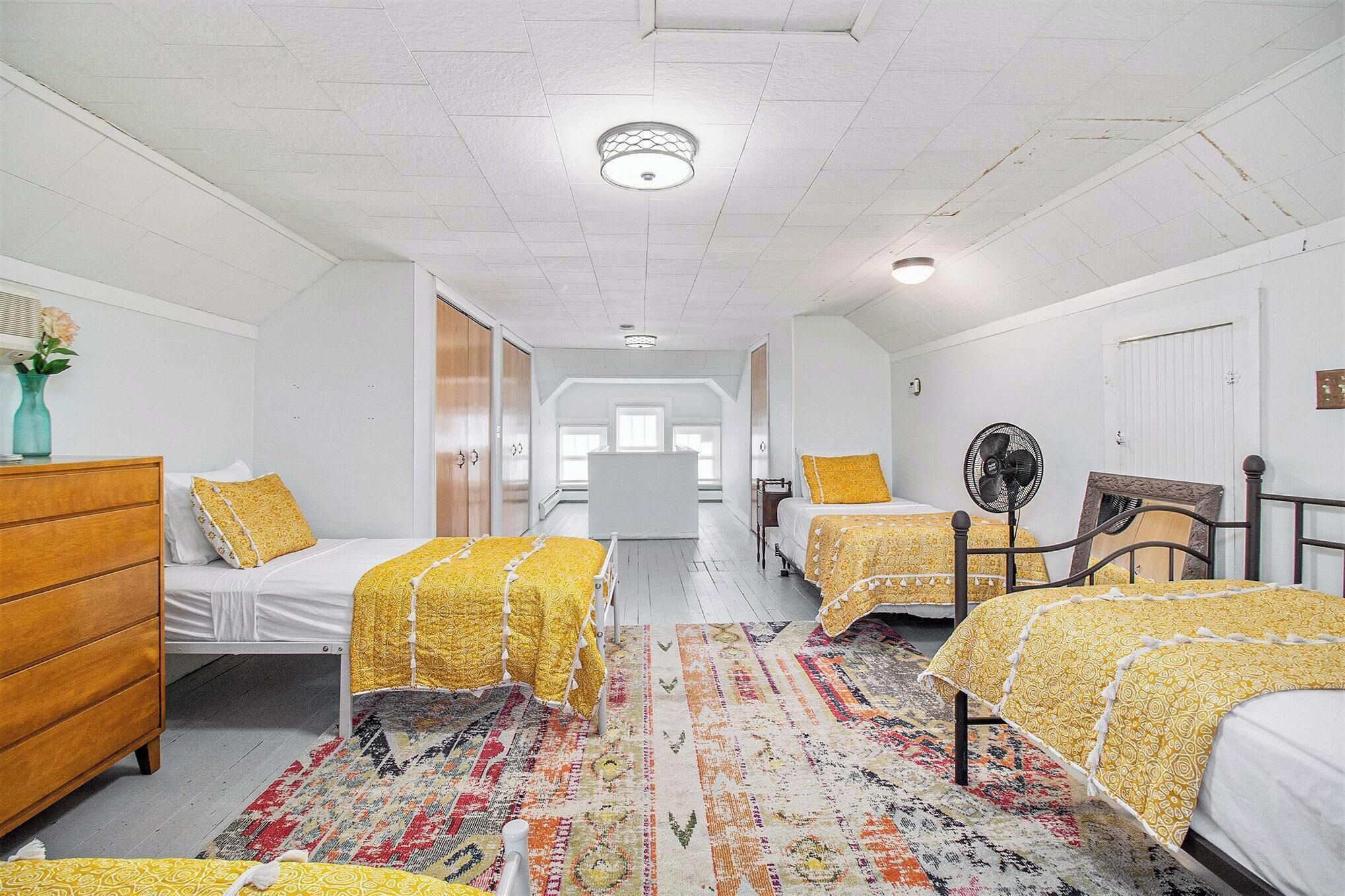 A room with two beds, a wooden dresser, a patterned rug, and a ceiling fan.