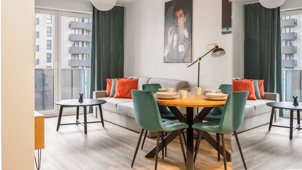 A modern living room with a dining area, featuring a sofa set, a round wooden table, and a large window with curtains. - LILY APART the Big Screen Apartment (Gdańsk)