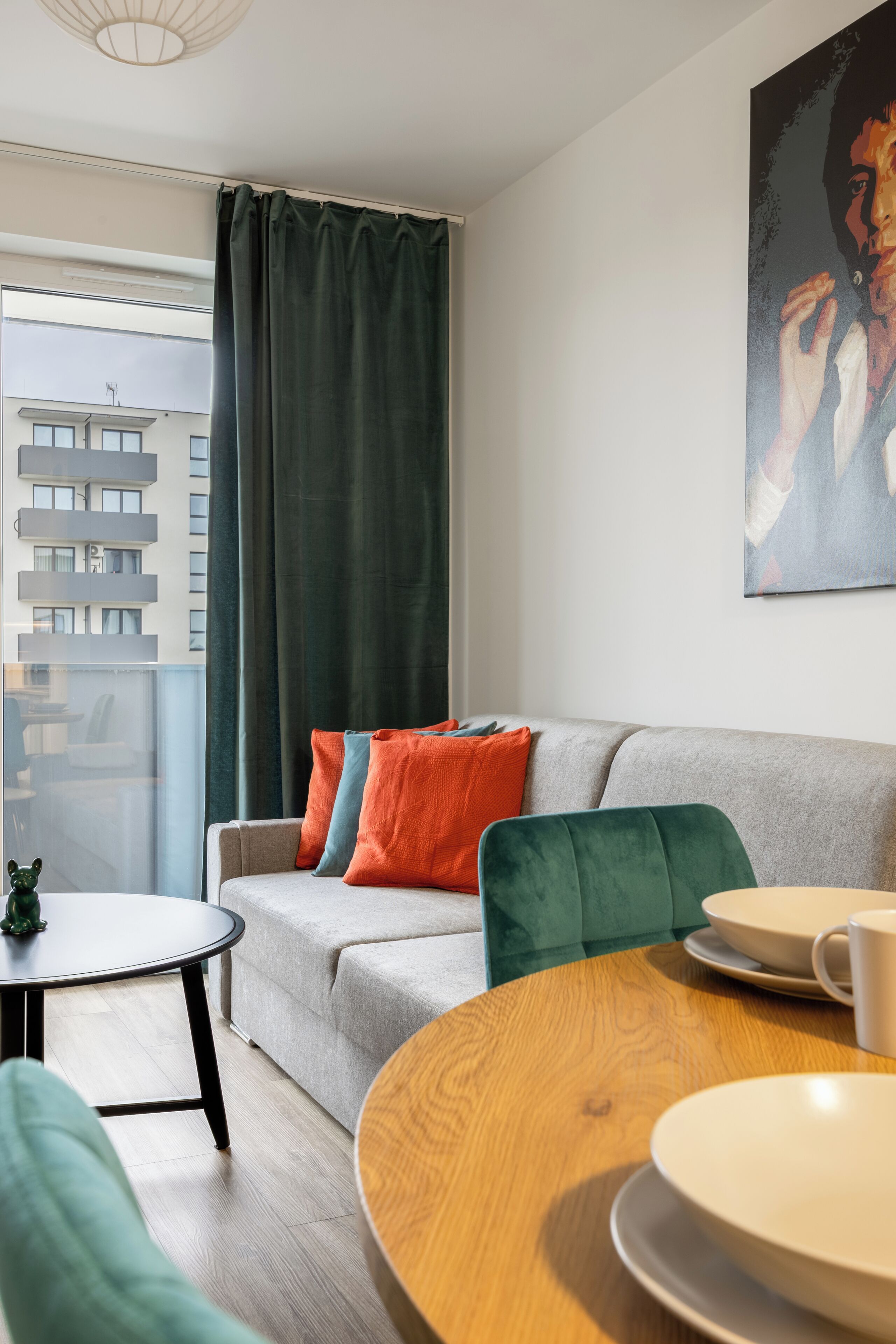 A modern living room with a grey sofa, colorful cushions, a round wooden table, and a view of a building through a window with a green curtain.