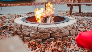 A stone fire pit with flames, surrounded by fallen leaves, with a picnic table and a hammock in the background. - Two Beach Homes with 2 Hot Tubs Sleeps 20+ (Michigan City)