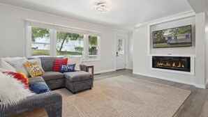 A modern living room with a sectional sofa, a fireplace, a flat-screen TV, and large windows. - Two Beach Homes with 2 Hot Tubs Sleeps 20+ (Michigan City)