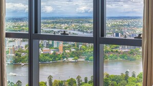Ein Zimmer mit einem großen Fenster und Blick auf eine Stadtlandschaft, einem Bett mit einer Türkis-farbenen Decke sowie einer Aussicht auf einen Fluss und Grünflächen.