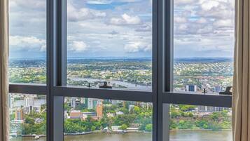 A room with a large window overlooking a cityscape, a bed with a teal blanket, and a view of a river and greenery.