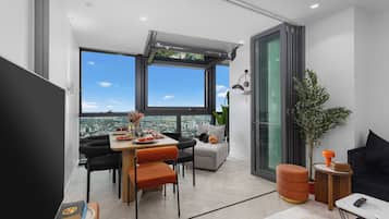 A modern living space with a dining area, seating, a sofa, a coffee table, and a large window offering a city view.