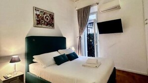 Cama bem arrumada com roupa de cama branca e azul escura, uma luminária de cabeceira, uma televisão na parede e uma janela com cortinas.