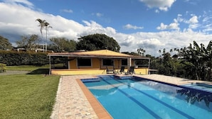 A swimming pool with a stone border, a yellow building with a tiled roof, and a grassy area with trees. - PARCEROS GROUP - 4-BEDROOM VACATION FARM, MORELIA, PEREIRA, COLOMBIA (La Estrella)