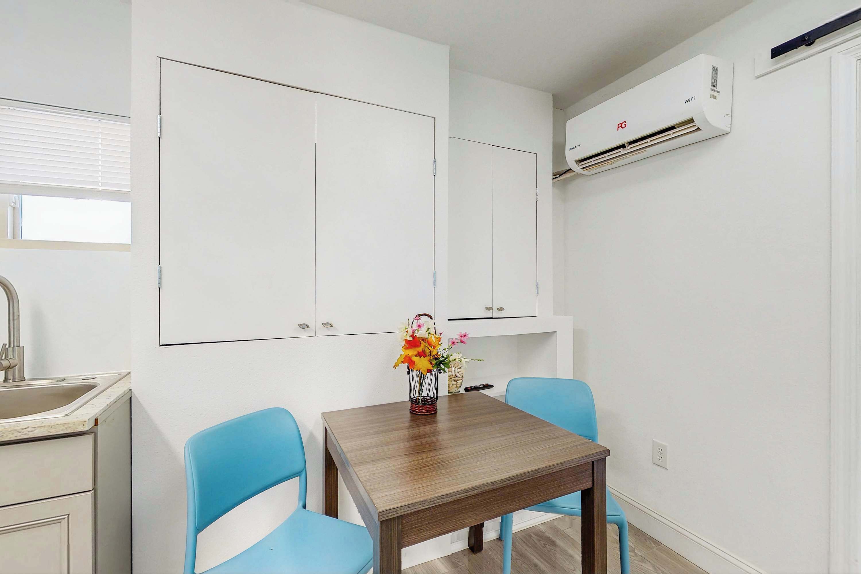 A small, clean room with a wooden table, two blue chairs, a white sink, and a white air conditioning unit.
