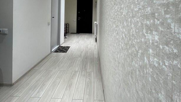 Ein Korridor mit hellem Holzboden, weißen Wänden und einer strukturierten Wand auf der rechten Seite.