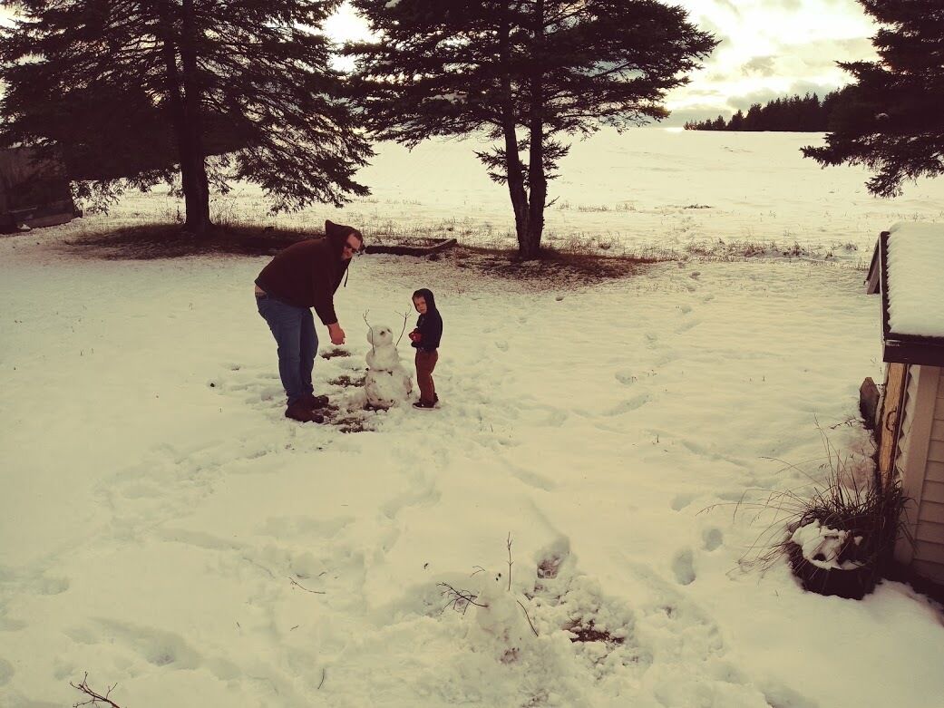 Une scène hivernale en plein air, avec de la neige, où l’on voit une personne et un enfant jouer dans la neige près d’une maison.