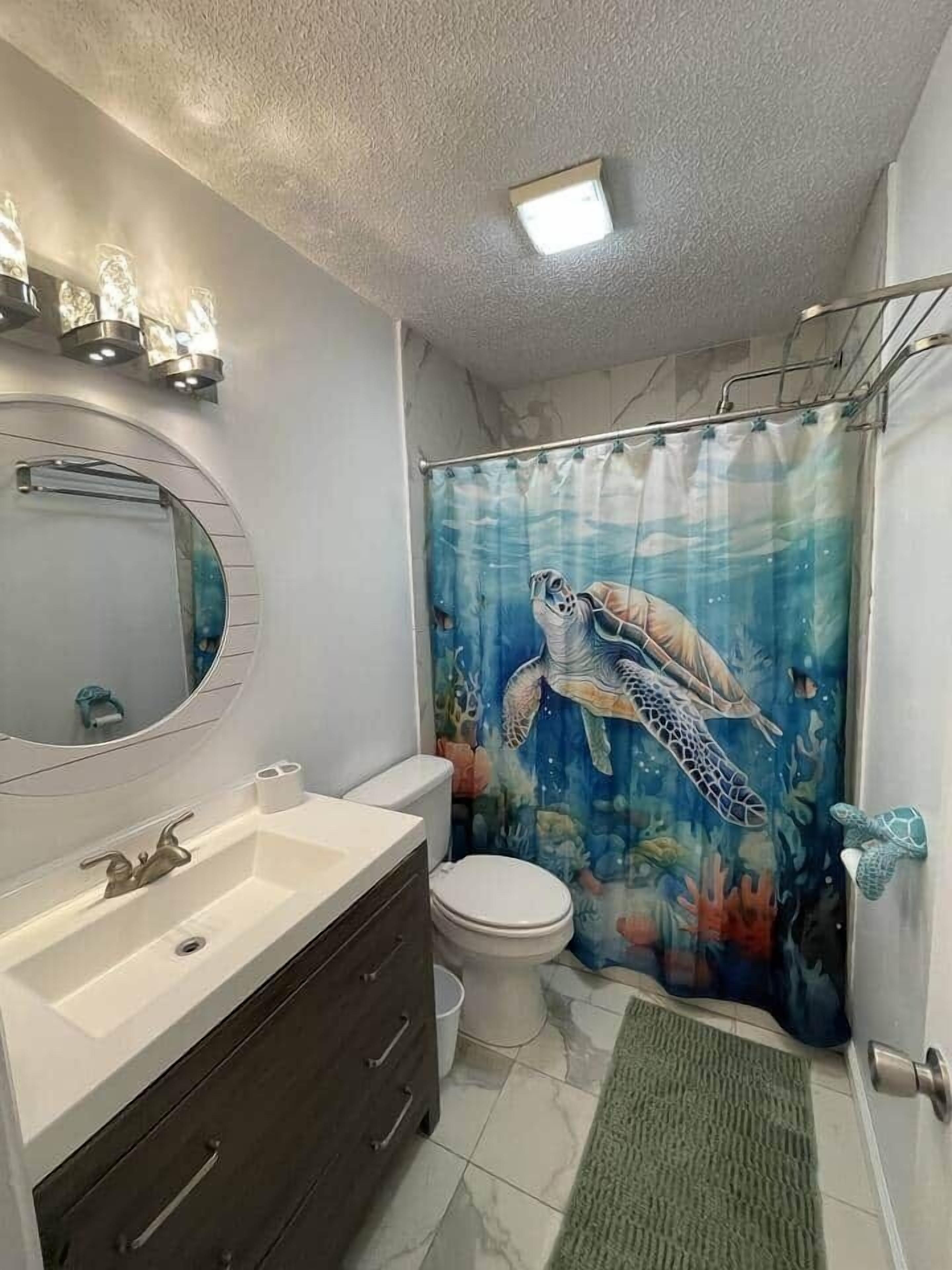 Une salle de bain avec un miroir rond, un lavabo, des toilettes et un rideau de douche orné d’un motif représentant une tortue de mer.