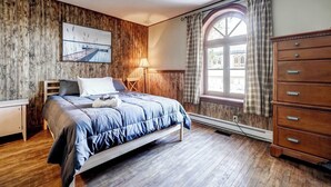 A bedroom with a wooden wall panel, a bed with a blue duvet, a wooden dresser, and a window with curtains. - The Historic Post Office, Le Bureau de Poste (Lac-des-Plages)