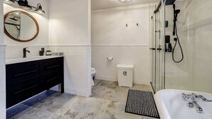 A modern bathroom with a black vanity, a round mirror, a glass shower enclosure, and a white sink. - The Historic Post Office, Le Bureau de Poste (Lac-des-Plages)