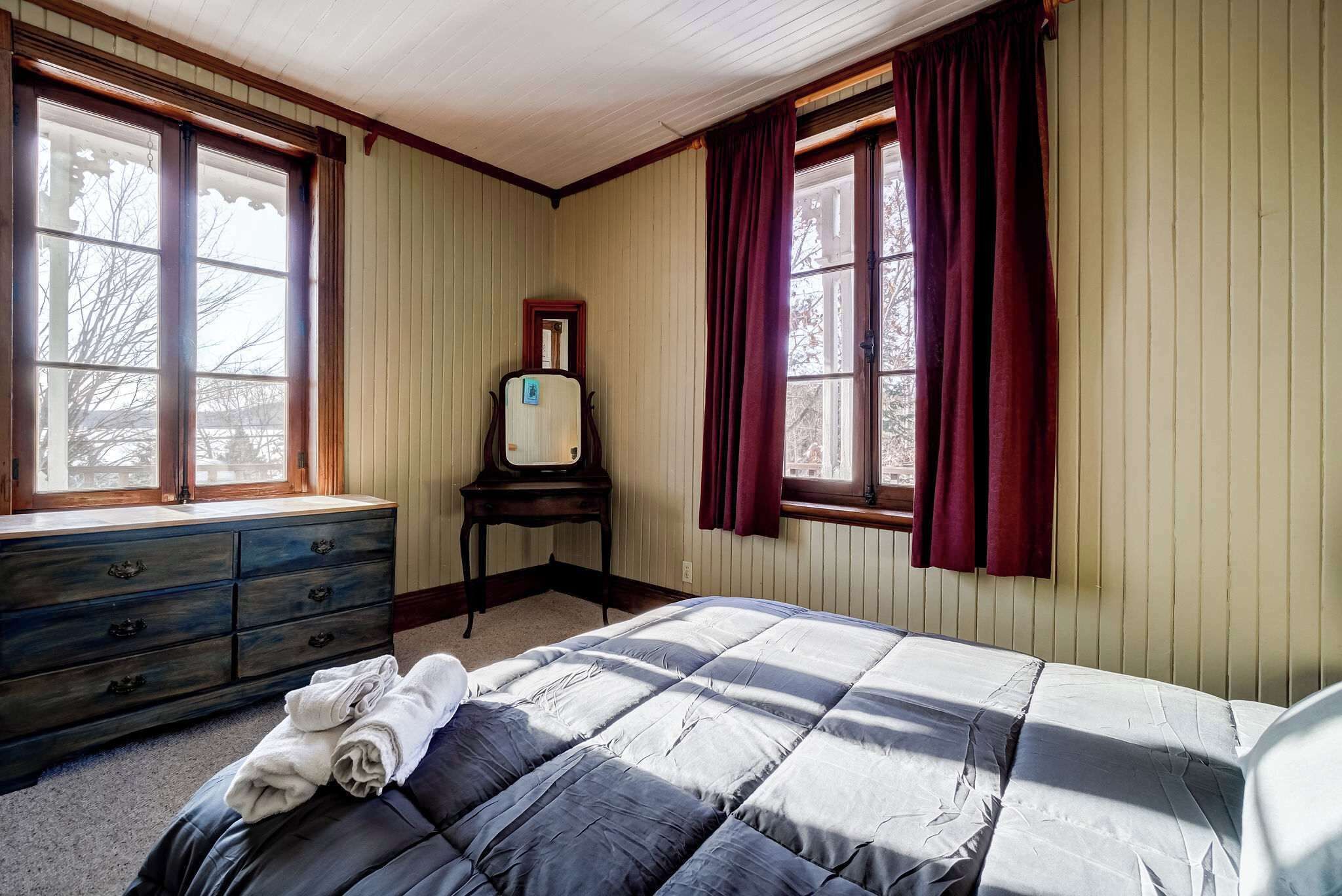 A bedroom with a bed, a dresser, a mirror, and two windows with curtains.