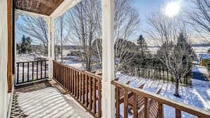 A covered porch with a wooden railing, a snow-covered ground, and leafless trees in the background. - The Historic Post Office, Le Bureau de Poste (Lac-des-Plages)
