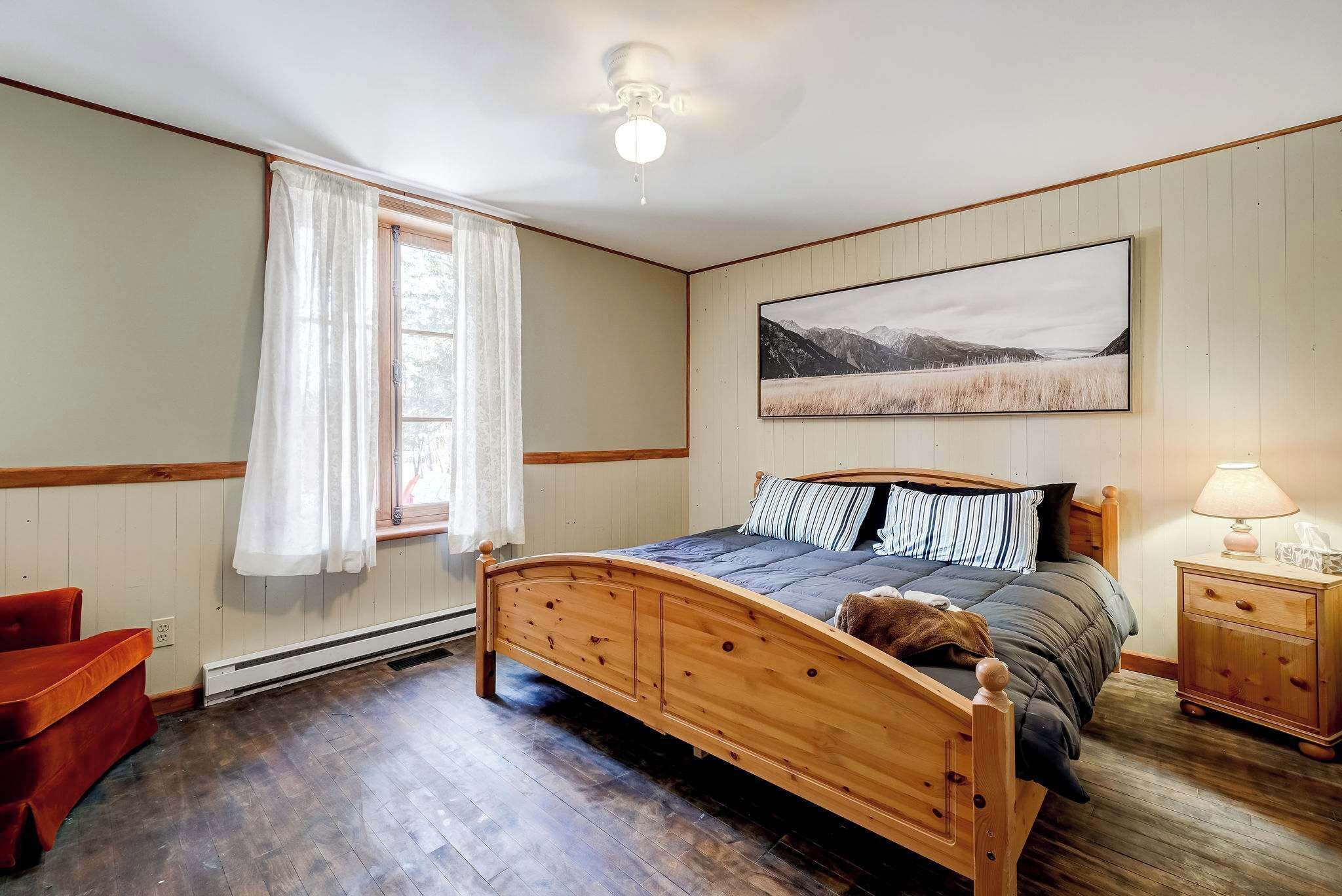 A bedroom with a wooden bed, a nightstand, a chair, a window with curtains, and a ceiling fan.