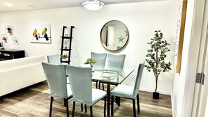 Et moderne spiseområde med et glasbord, grå polstrede stole, trægulv og en dekorativ plante.