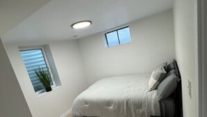A bedroom with a bed, a small window, a potted plant, and a nightstand. - A Sleek Modern 4-bedroom house with updated amenities in welcoming Westminster (Westminster)