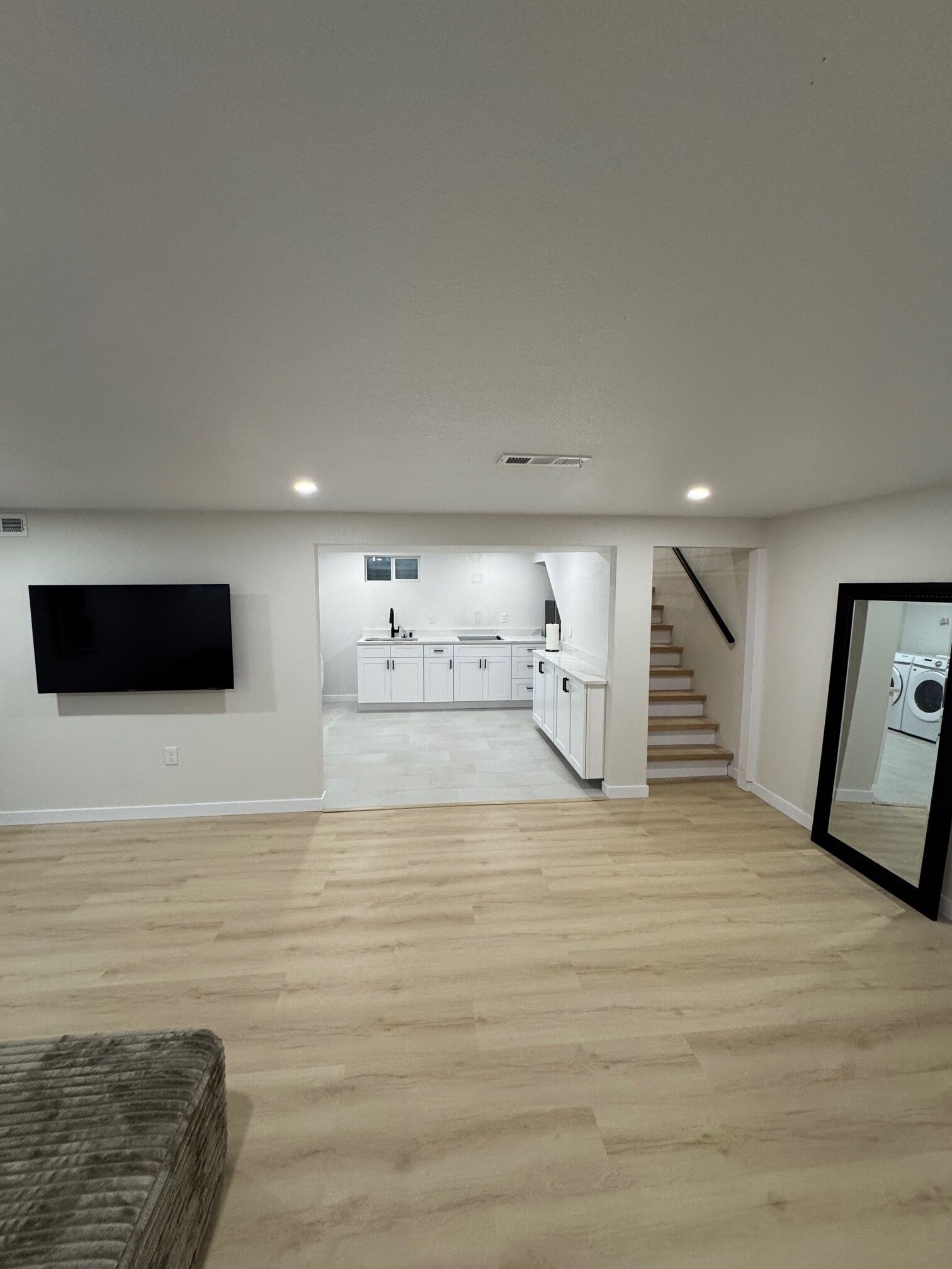 A spacious living area with a flat-screen TV mounted on the wall, a kitchen in the background, and a staircase leading to another level.
