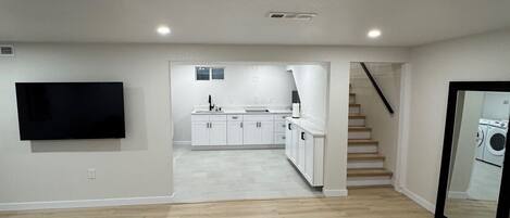 A spacious living area with a flat-screen TV mounted on the wall, a kitchen in the background, and a staircase leading to another level.