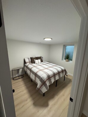 Ein Schlafzimmer mit Bett, kleinem Tisch, Fenster mit Pflanze und Deckenleuchte.