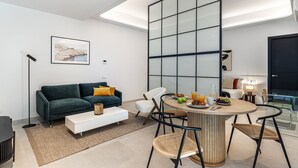 A modern living room with a sofa, coffee table, and dining area. There is a large window with a view, a painting on the wall, and a lamp. - Waou Urban Home Salamanca V (Madrid)