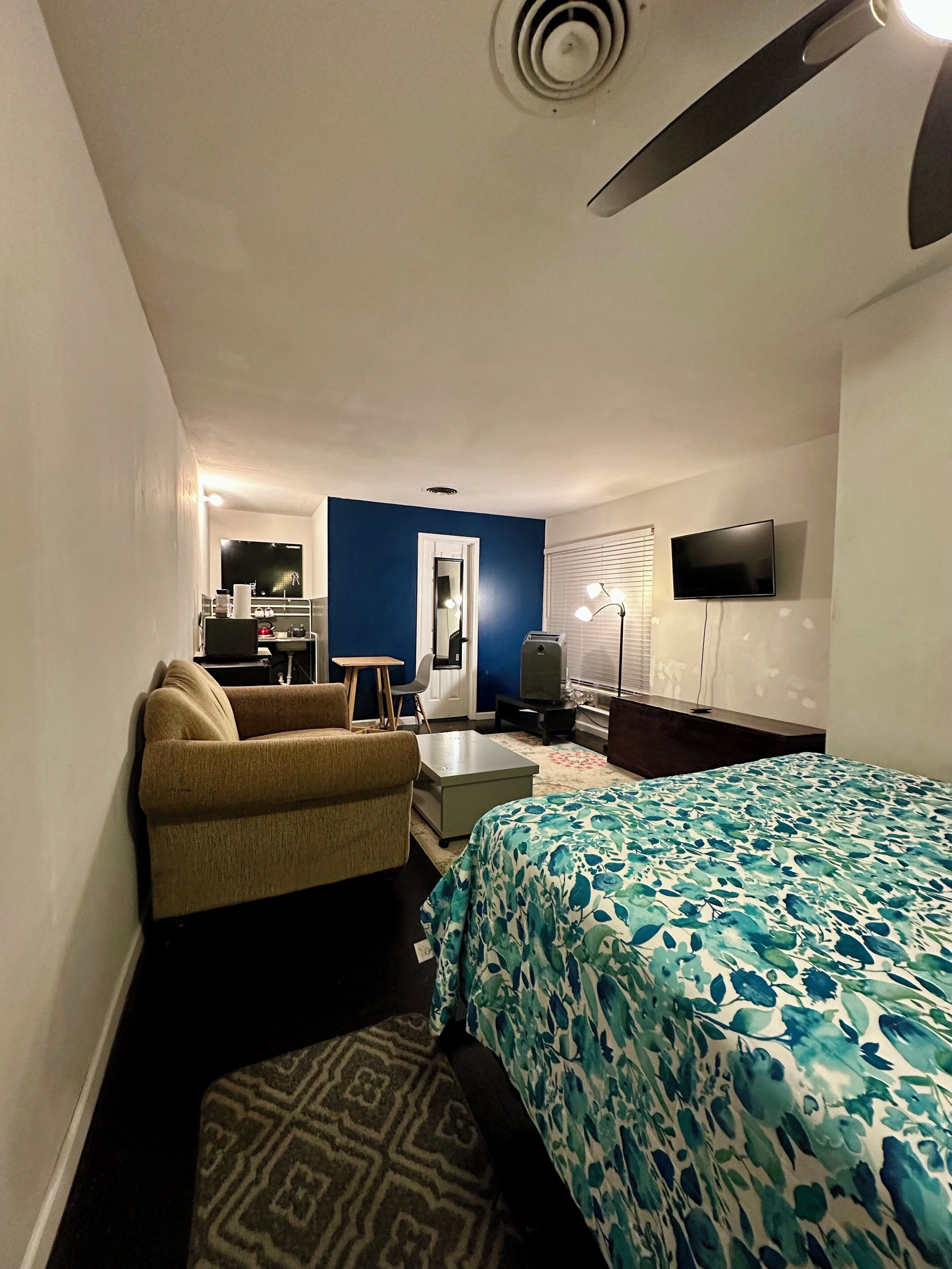 Quarto de hotel com cama, sofá, escrivaninha, televisão e ventoinha no teto.