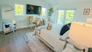 A bedroom with a bed, a desk, a chair, a television, a ceiling fan, and a balcony with a view of greenery.