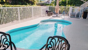 A pool area with a glass table and metal chairs, surrounded by a white fence and palm trees.