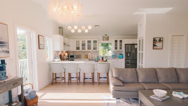 A modern kitchen with white cabinets, a central island with bar stools, a dining area with a sofa, and a chandelier.