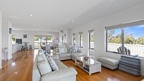Amplia sala de estar con un sofá en forma de L, una mesa de centro y un comedor con mesa y sillas. Grandes ventanas ofrecen luz natural y vistas al exterior.