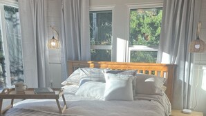 Ein Schlafzimmer mit einem hölzernen Bett, weißer Bettwäsche, einem Nachttisch mit Lampe und Blick auf Grün durch das Fenster.