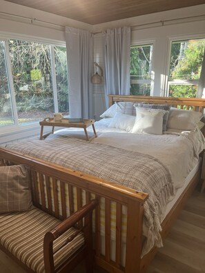 Un dormitorio con una cama grande, un banco de madera y vistas a zonas verdes por las ventanas.