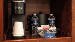 Un mini-bar comprenant une machine à café Keurig, des bouteilles d’eau, une tasse blanche et une paire de verres.