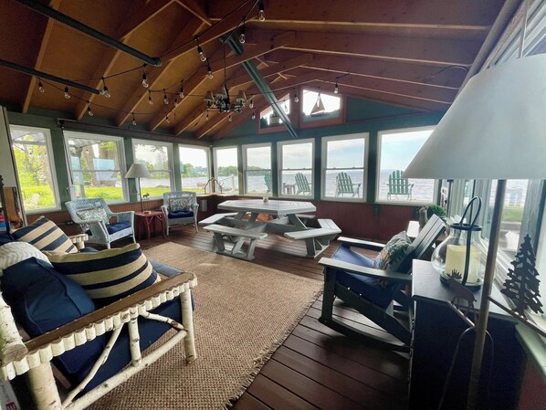 A covered deck with wooden flooring, outdoor seating, and large windows overlooking a body of water.