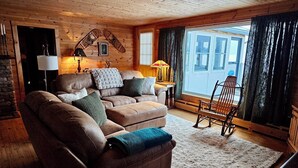 A cozy living room with a leather sofa, a wooden ceiling, and a large window with curtains. - 130 Feet of Lakefront with Unforgettable Views and Access (Plattsburgh)