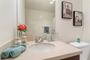 Une salle de bain avec un lavabo beige, deux tableaux encadrés au mur, des toilettes et un récipient vert sur le côté.