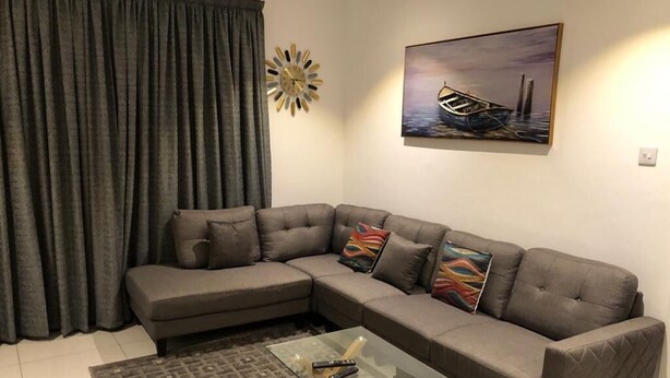 Ein Wohnzimmer mit einem Ecksofa, einem Glastisch, einem gemusterten Teppich, einem Bild eines Bootes und einer Dekotischuhr.