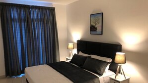 Ein Schlafzimmer mit einem Bett, zwei Nachttischlampen, einem Nachttisch, einem gerahmten Bild an der Wand und einem Fenster mit Vorhängen.