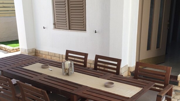An outdoor dining area with a long wooden table and chairs, a tiled floor, and a white wall with a window and shutter. - Marvelous 5-bedroom house with great terrace in Pješčana Uvala (Pješčana Uvala)