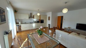 Una cucina moderna con mobili bianchi, un'area pranzo con tavolo in vetro e un soggiorno con divano e televisore.