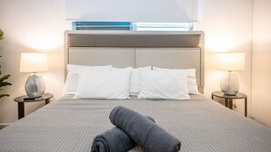 Ein ordentlich bezogenes Bett mit grauer Decke, zwei zusammengerollten Handtüchern, zwei weißen Kissen und je einer Nachttischlampe auf jeder Seite.