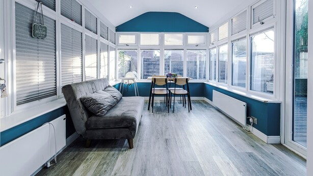 A room with a grey sofa, a small table with chairs, and large windows with blinds. - Modern 3-Bed - Near Etihad & City Centre (Manchester)