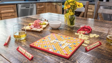 A wooden table with a Scrabble board, a bowl, a glass of beer, and a plate of food.