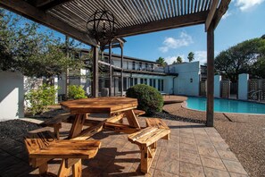 A covered outdoor area with picnic tables, a pool, and a building in the background.