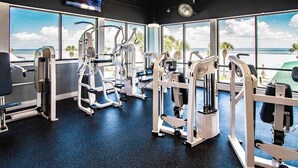 Ett gym med olika träningsmaskiner, stora fönster med utsikt över stranden och en klocka på väggen.