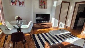 A modern living room with a glass dining table, white chairs, a brown sofa, and a TV on a wooden stand.