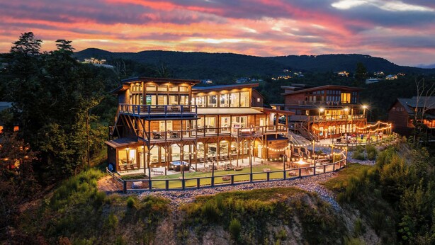 A multi-story wooden lodge with a deck and outdoor seating area, surrounded by forested hills. - 6BR for 26 | Pool, Hot Tub, Game Loft, & Mini Golf (Sevierville)